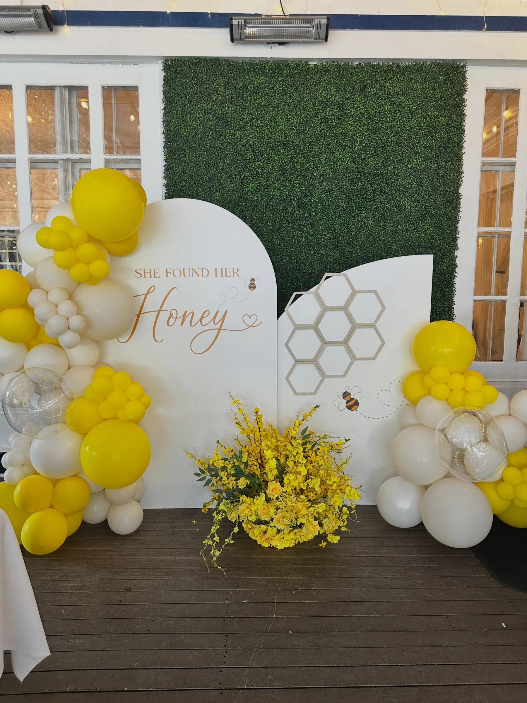 bridal shower backdrop rental in Naperville — white arched panels with gold 'She Found Her Honey' lettering, honeycomb detail, green hedge wall and yellow balloon garlands at Mesón Sabika