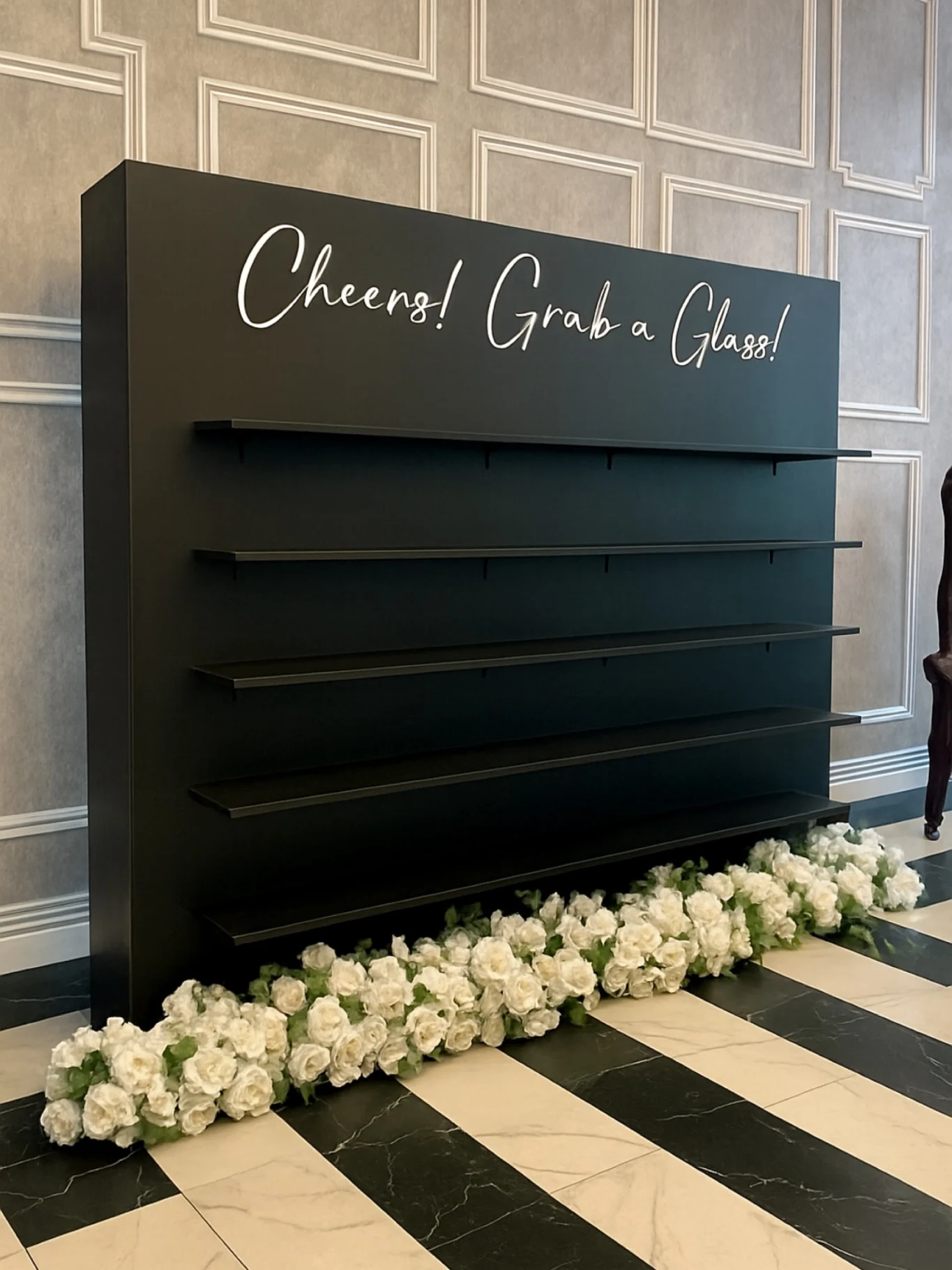 Champagne wall rental in Chicago with a black five shelf wall white script Cheers Grab a Glass lettering and white roses at its base