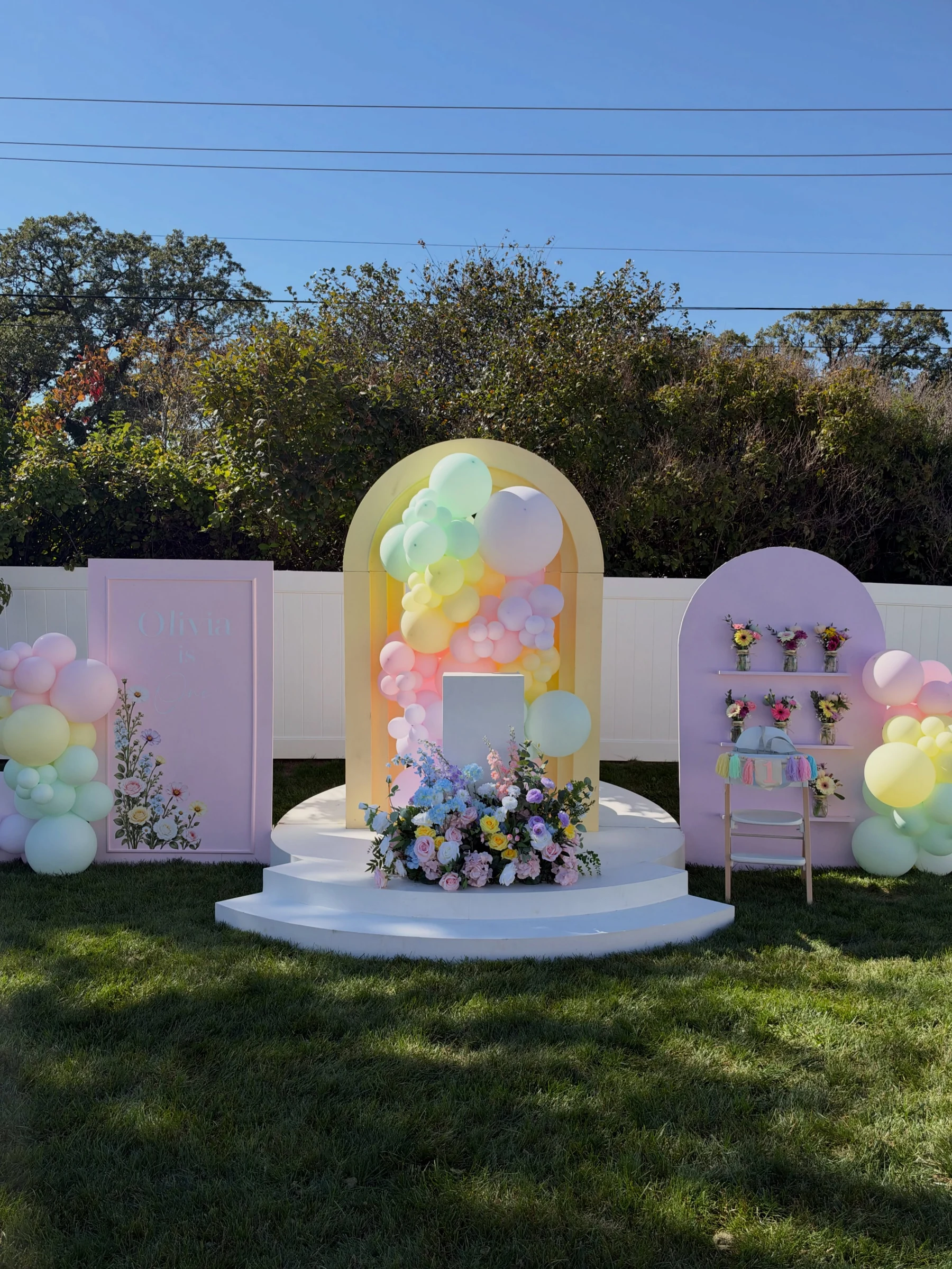 first birthday backdrop rental in Chicago — yellow arch filled with pastel balloons, pink Olivia is One panel and lavender flower-shelf panel on a white platform in a backyard