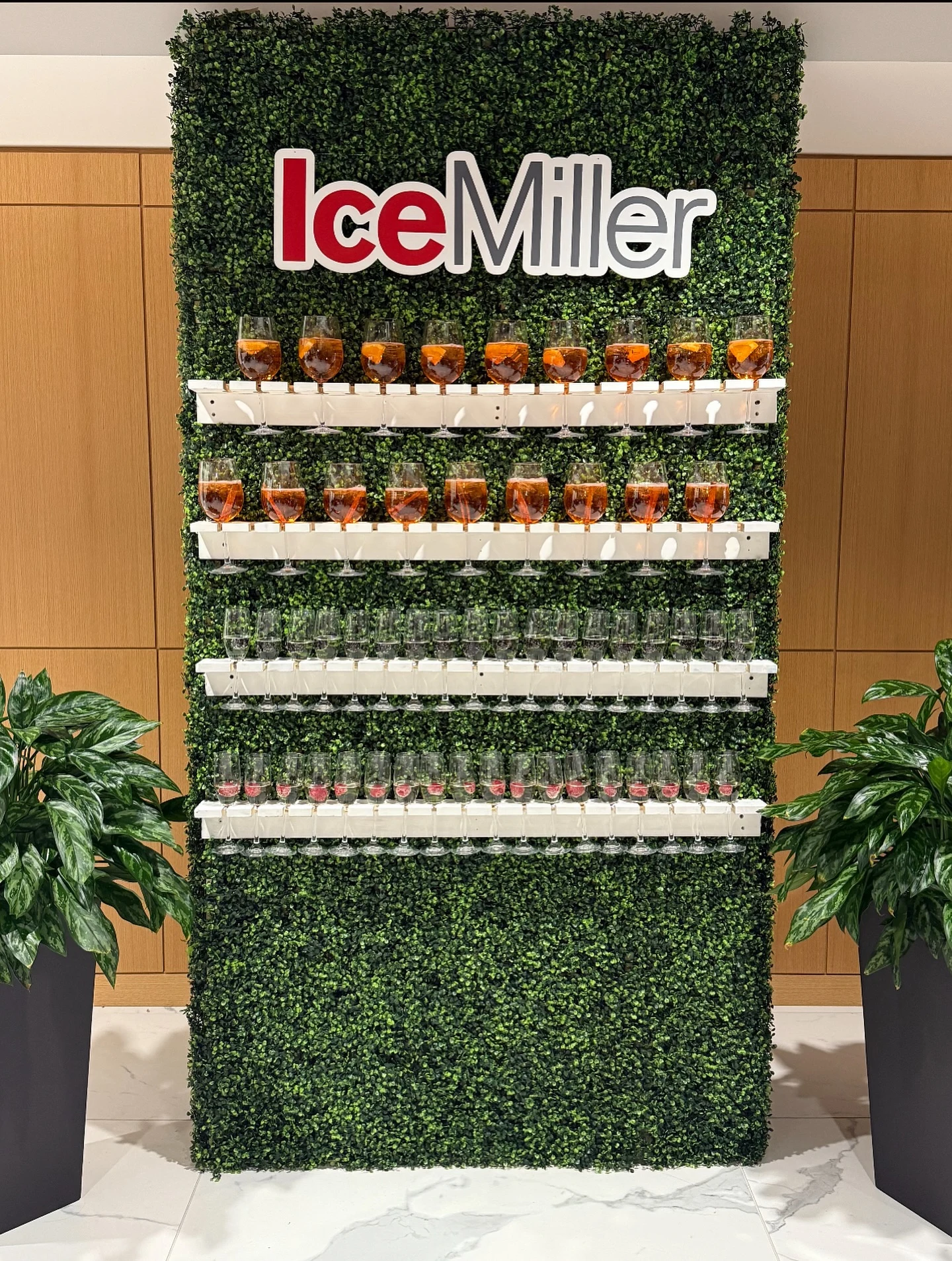Champagne wall rental in Chicago with a half boxwood champagne wall holding four shelves of drinks under a custom Ice Miller logo sign in an office lobby