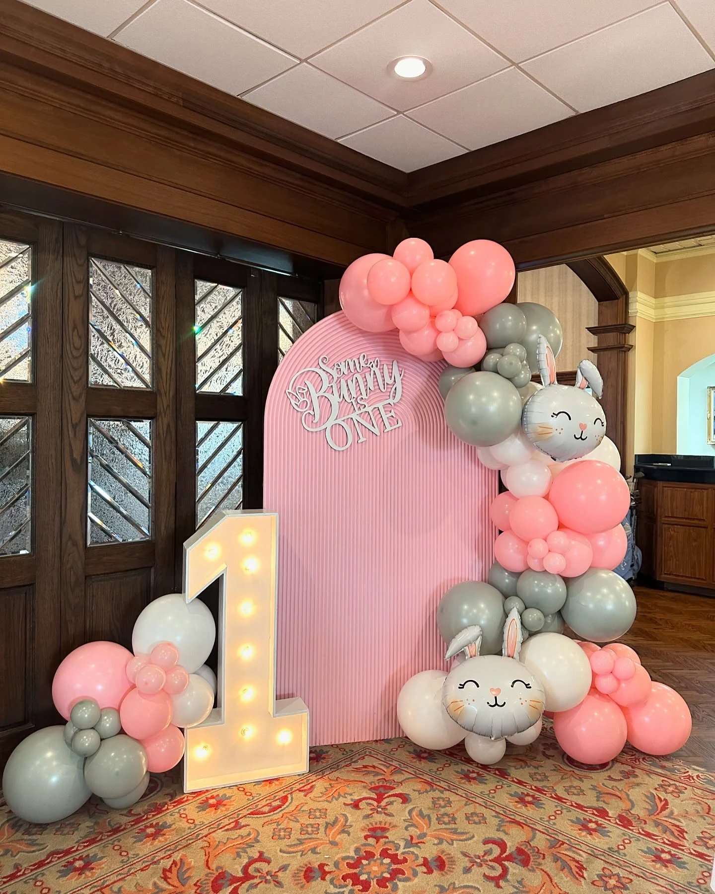 First birthday backdrop rental in Lemont — a pink ribbed arch panel lettered 'Some Bunny is ONE' at Cog Hill, framed by a coral pink, sage and white balloon garland with two grey bunny-head foil balloons and a light-up marquee number 1 at its base
