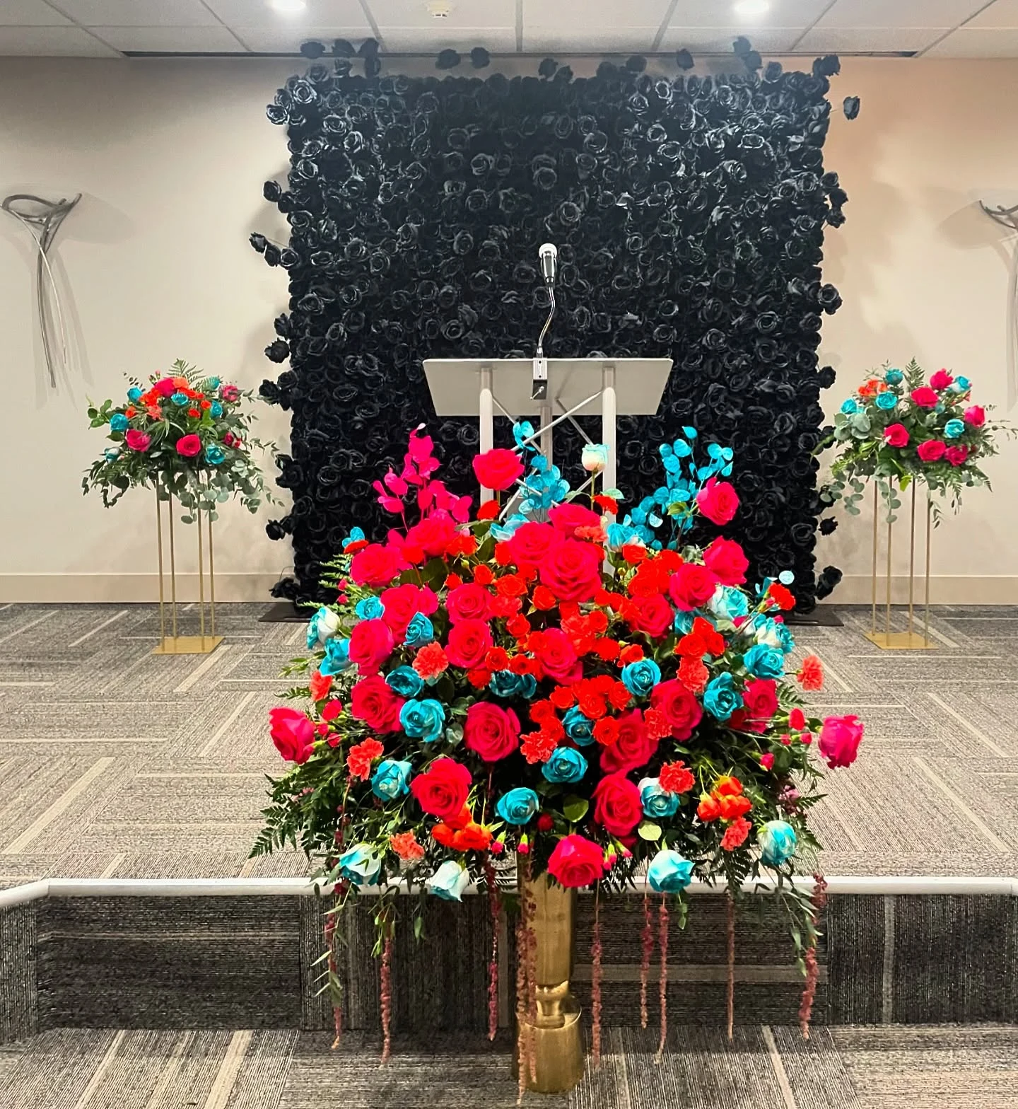 Flower wall rental in Chicago — black rose flower wall behind a podium on a stage, flanked by tall pink, red and turquoise floral arrangements
