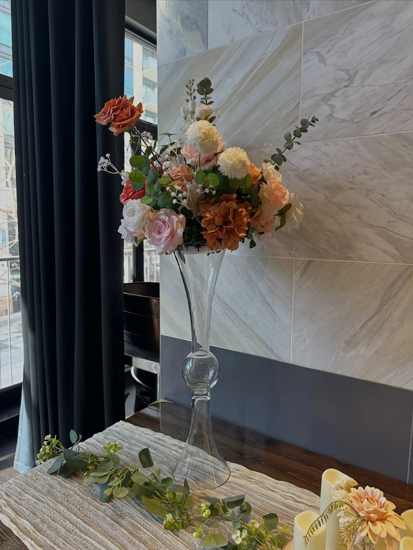 Matching wedding centerpiece in Chicago with a tall glass trumpet vase of peach pink white and rust roses above a gauze runner eucalyptus garland and pillar candles