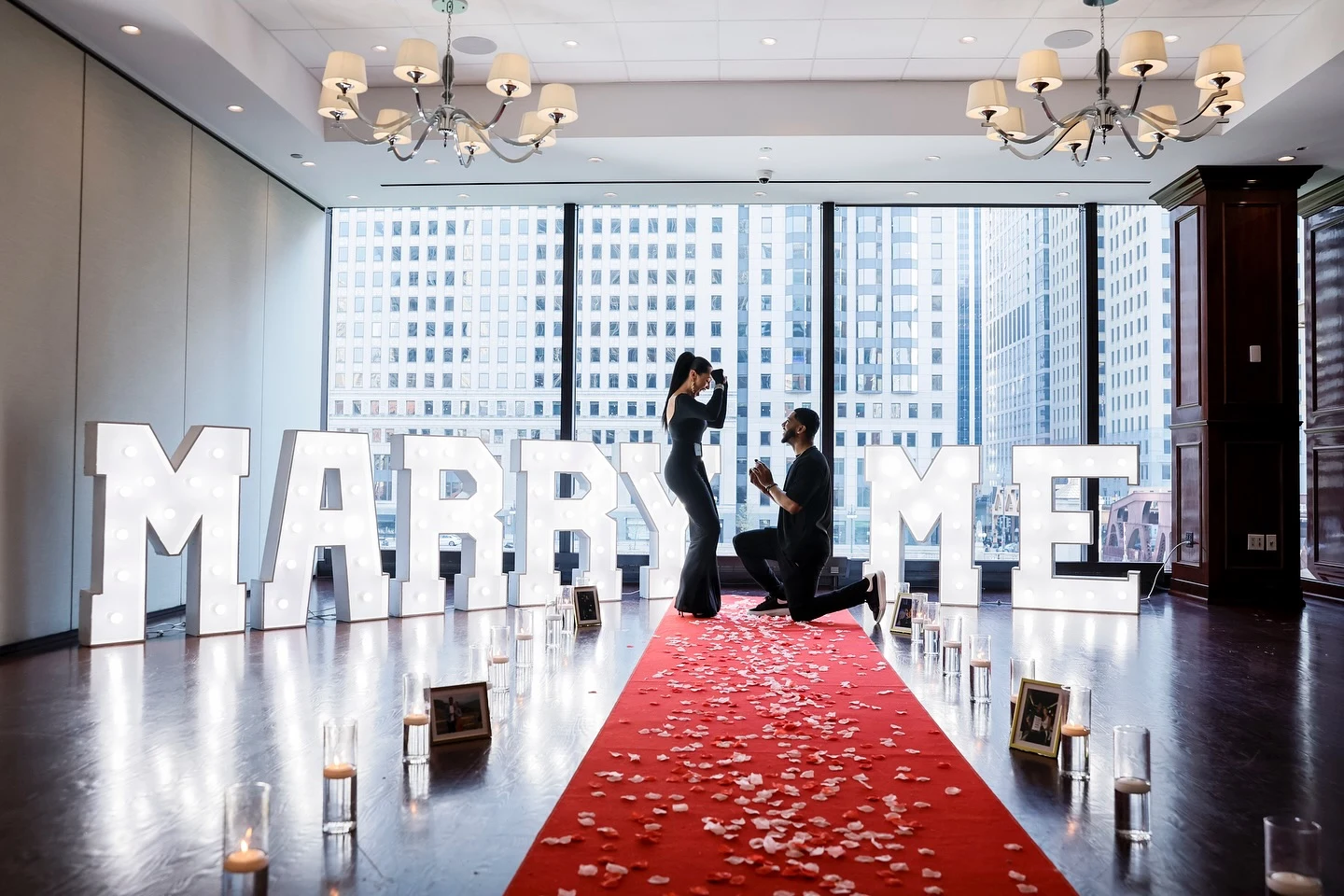 Proposal rental in Chicago — giant white MARRY ME marquee letters behind a red petal aisle as a man proposes on one knee in front of downtown windows at Chicago Cut Steakhouse