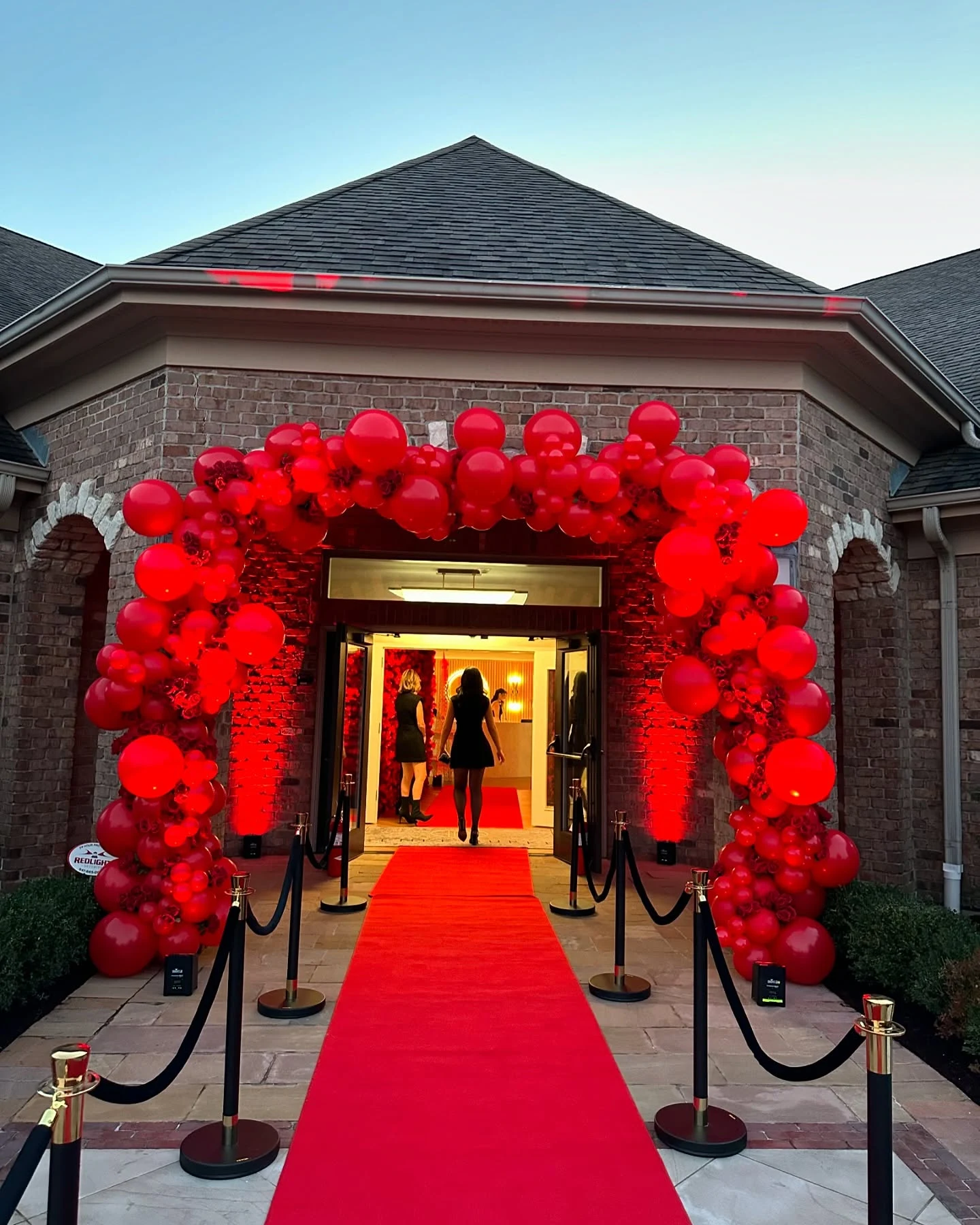 Balloon garland rental in South Barrington — all-red balloon garland with roses woven through it framing a brick entrance over a red carpet at dusk