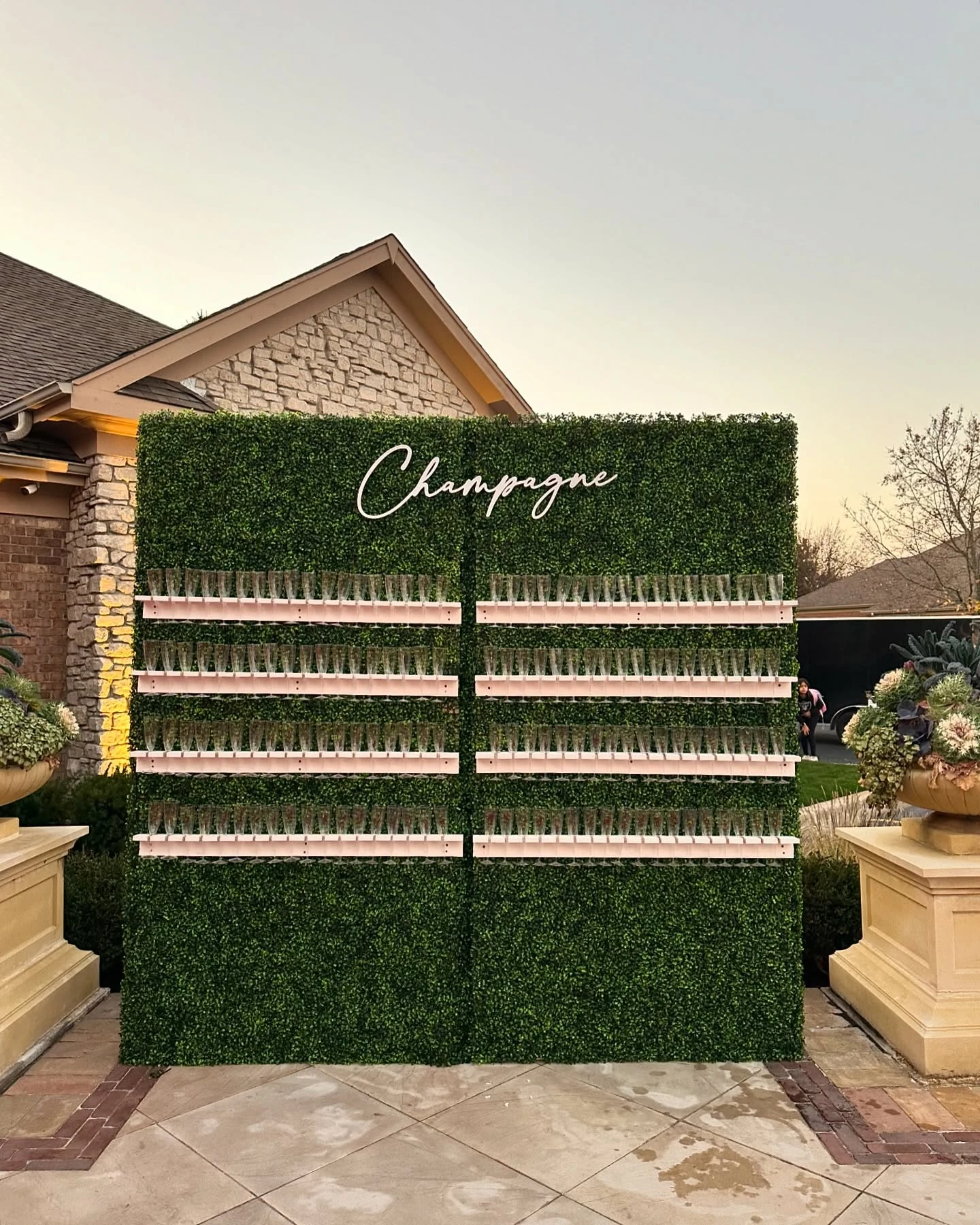 Champagne wall rental in South Barrington — full boxwood champagne wall with eight shelves of flutes and a script Champagne sign outdoors at dusk