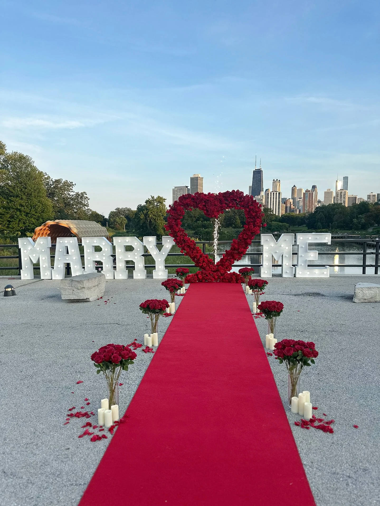 Heart arch rental in Chicago — red rose heart arch flanked by light-up MARRY ME marquee letters, a red carpet aisle lined with rose bouquets and candles, and the skyline over Lincoln Park's South Pond