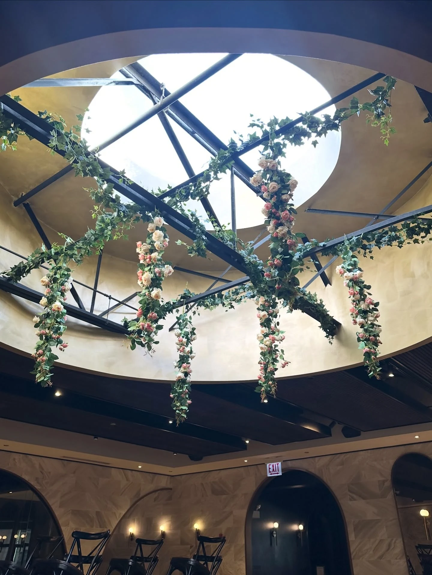 Hanging ceiling florals in Chicago with pink and cream rose garlands and ivy draped from black beams beneath a circular skylight at Chicago Winery