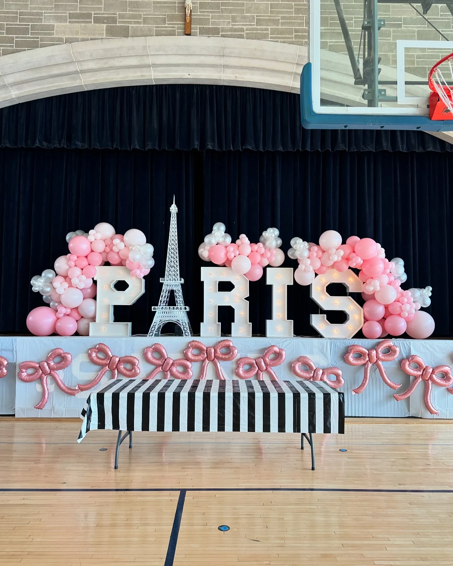 Paris marquee letter backdrop rental in Chicago — illuminated letters spelling PARIS with a white Eiffel Tower model standing in for the A, pink and pearl balloon garlands and rose-pink bow balloons on a school gym stage at Queen of All Saints