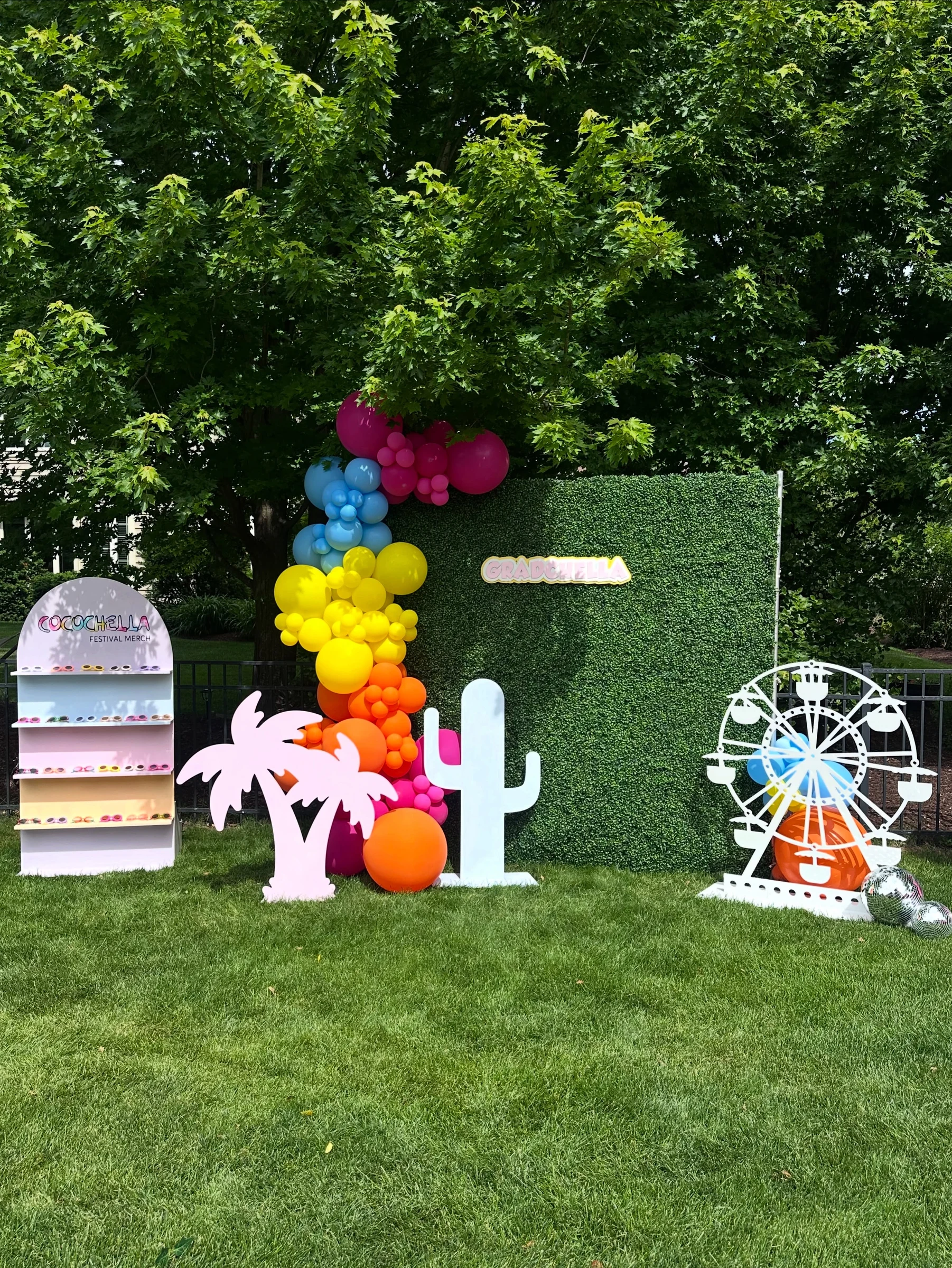 graduation party hedge wall backdrop rental in Batavia — green hedge wall with pink Gradchella sign, rainbow balloon garland, palm tree, cactus and Ferris wheel cutouts on a backyard lawn