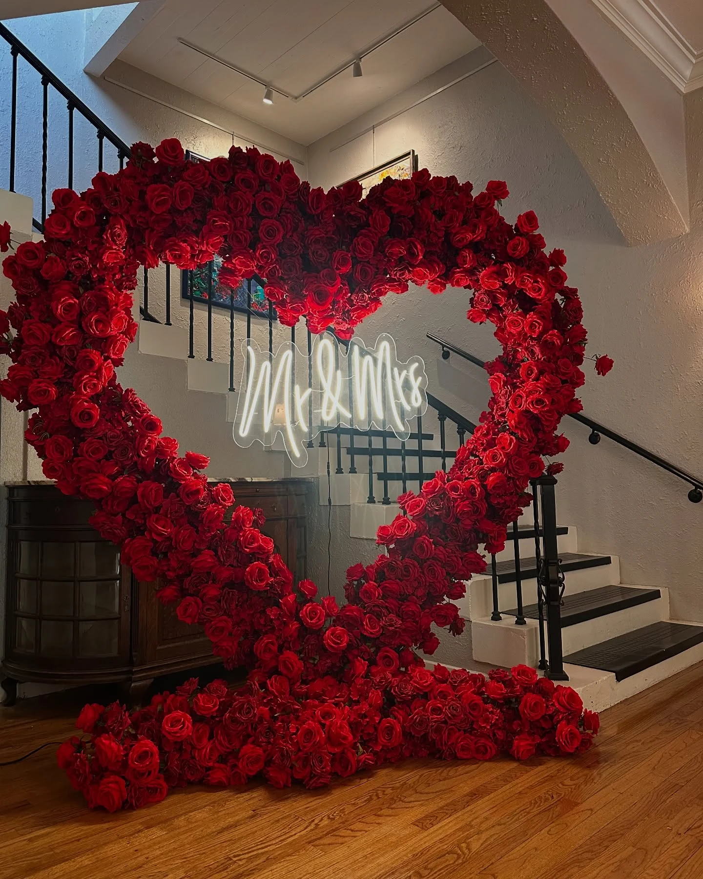 Heart arch rental in Chicago — red rose heart arch with a white Mr and Mrs neon sign at the foot of an indoor staircase, roses pooling on a wood floor
