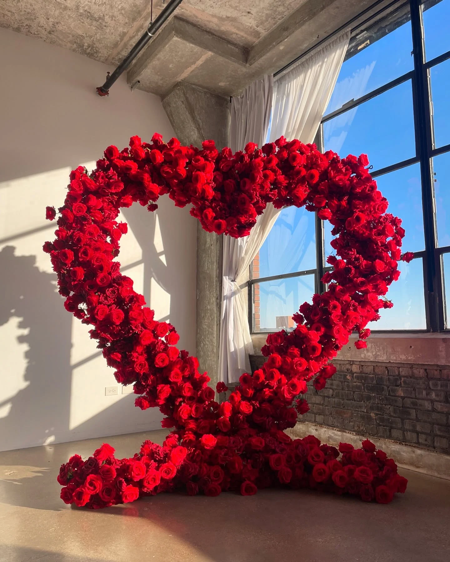Heart arch rental in Chicago — red rose heart arch standing alone in a sunlit loft studio with industrial windows, roses pooling at its base