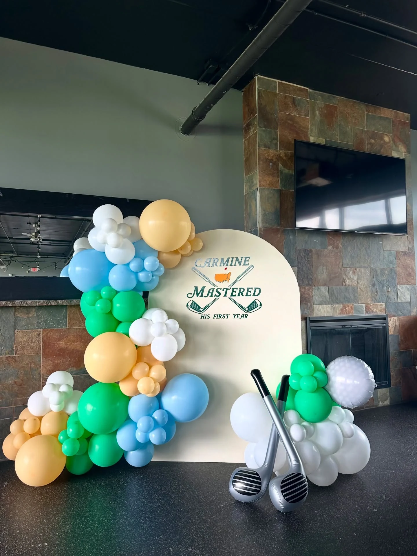 golf first birthday backdrop rental in Wood Dale — a cream arch panel lettered Carmine Mastered His First Year with crossed golf clubs, a green blue white and peach balloon garland, and inflatable golf club balloons at 390 Golf Experience