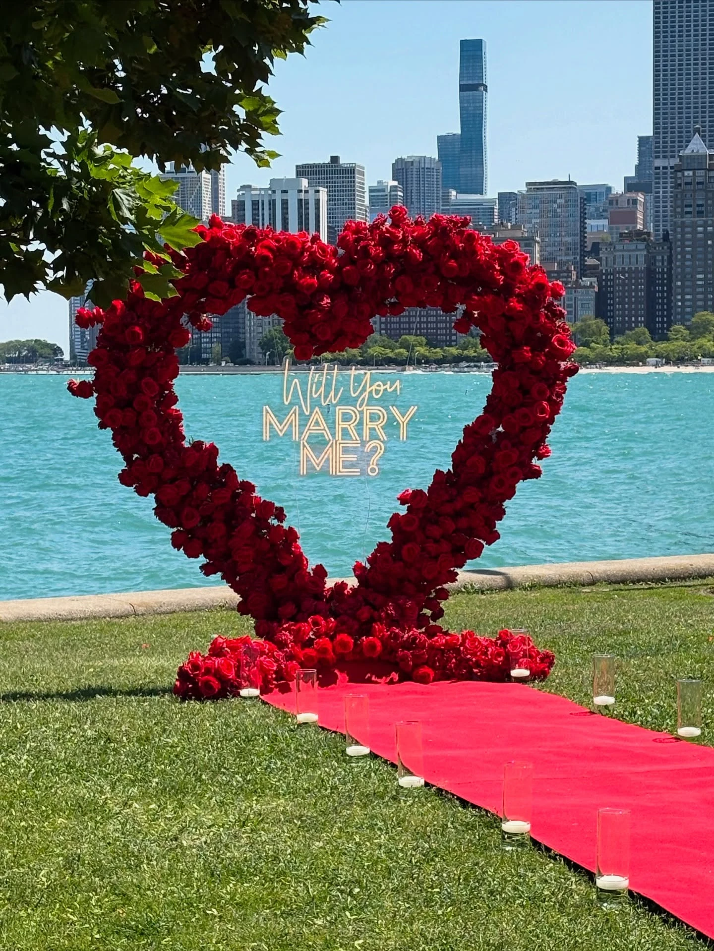 Heart arch rental in Chicago — red rose heart arch with a Will You Marry Me neon sign, a red carpet aisle and candle cylinders on grass at North Avenue Beach with Lake Michigan and the skyline behind