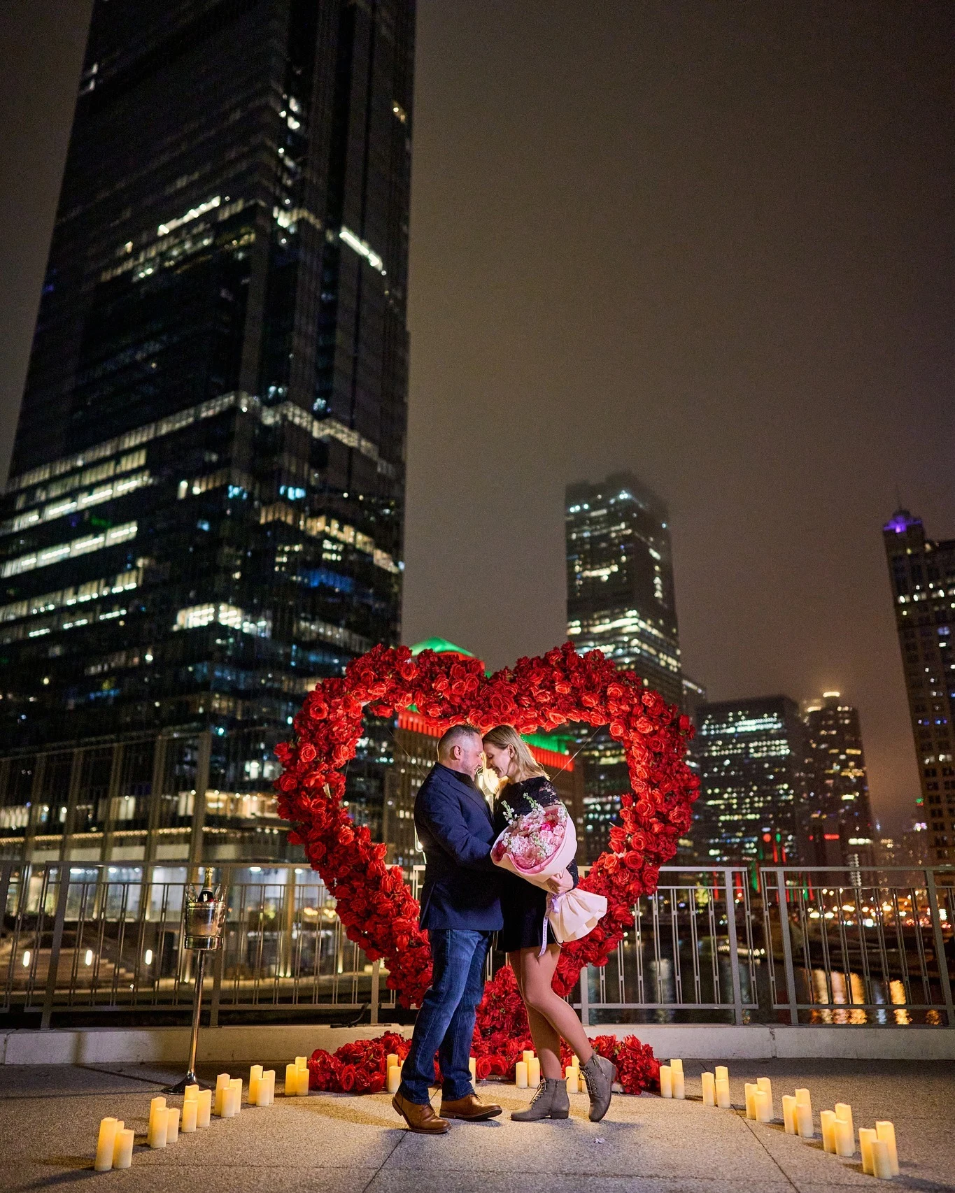 Red rose heart arch in Chicago at night with a couple forehead to forehead the woman holding a pink and white bouquet beside a champagne bucket