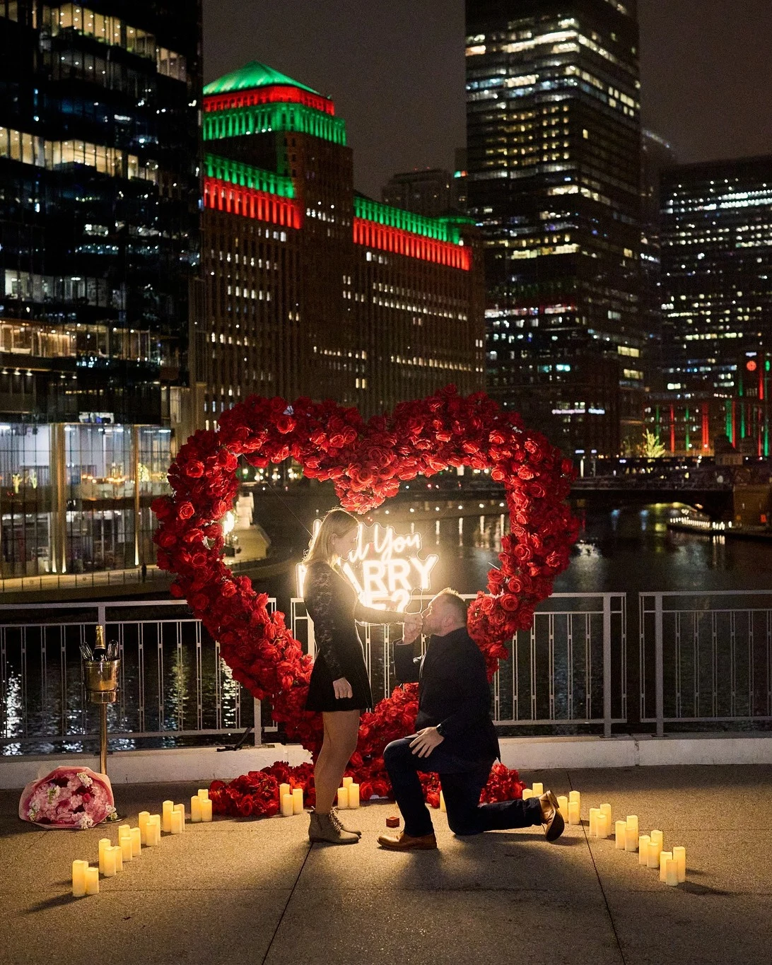 Heart arch rental in Chicago with a red rose heart arch and neon Will You Marry Me sign as a man kneels inside a heart of candles at River Point Park
