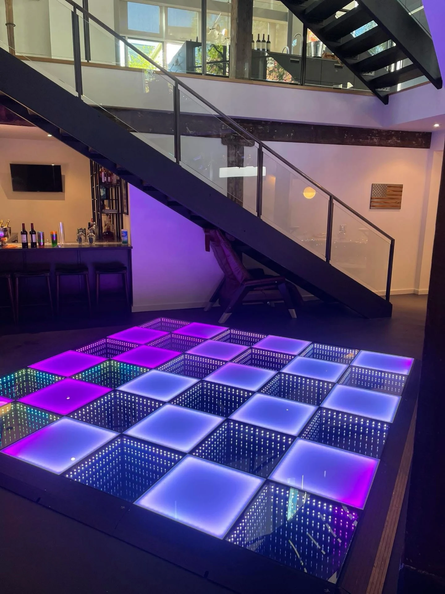 LED dance floor rental in Chicago with magenta and cyan infinity panels glowing in a modern home beside a glass-railed staircase and home bar.