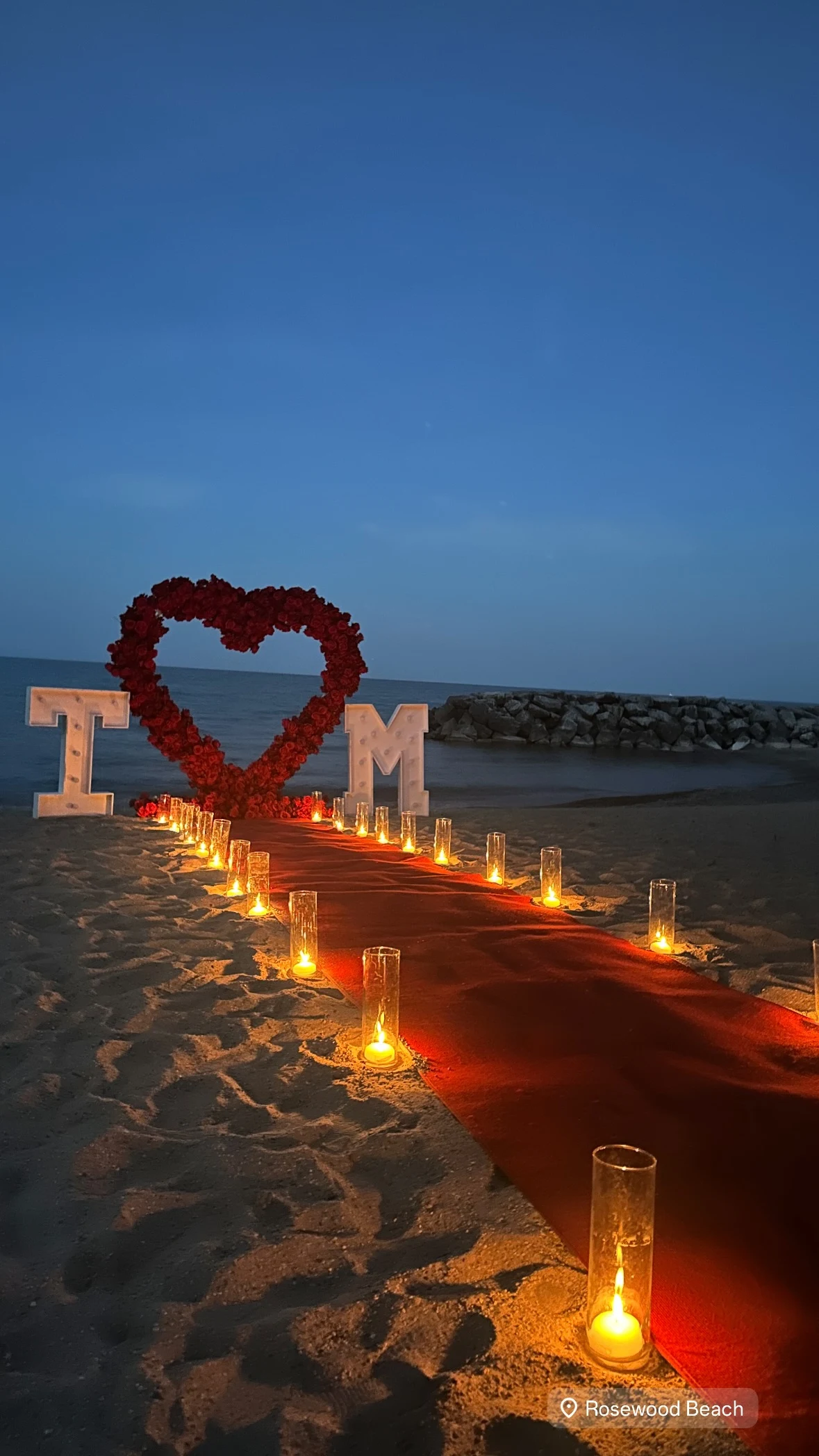 Heart arch rental in Highland Park — red rose heart arch on the sand at Rosewood Beach at dusk with a candle-lined red aisle and marquee initial letters