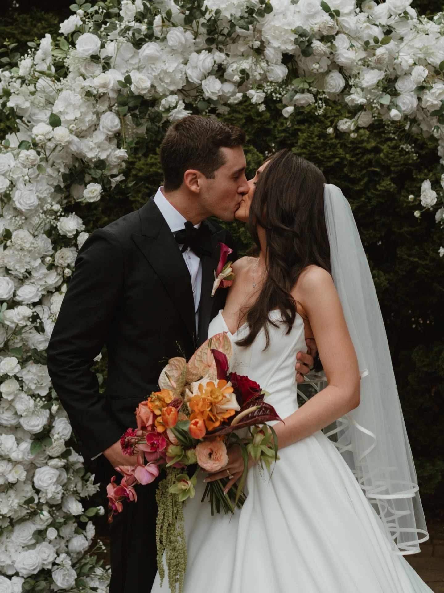 Wedding arch rental in Chicago — couple kissing in close-up in front of a dense white rose floral arch, bride holding an orange and pink bouquet
