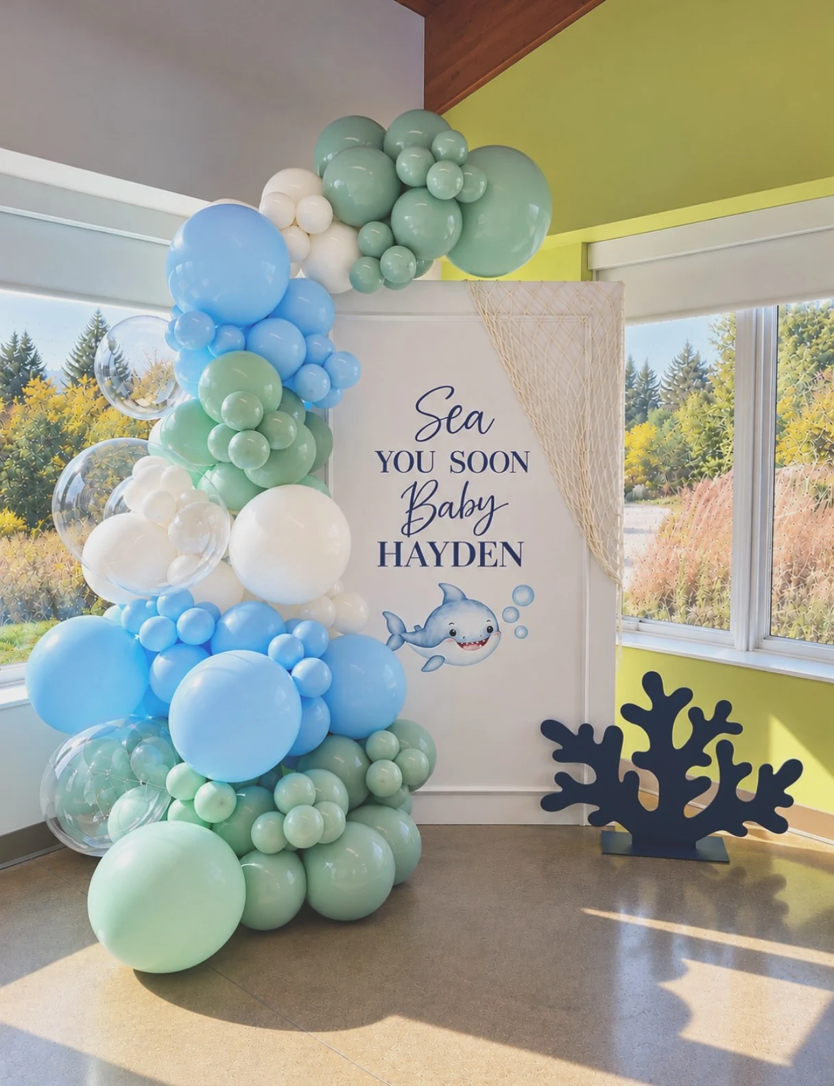 Baby shower backdrop rental in Chicago — a white 'Sea You Soon' backdrop panel lettered for Baby Hayden with a baby shark illustration and fishing-net drape, framed by a seafoam green, sky blue and white balloon garland with clear bubble balloons and a navy coral cutout