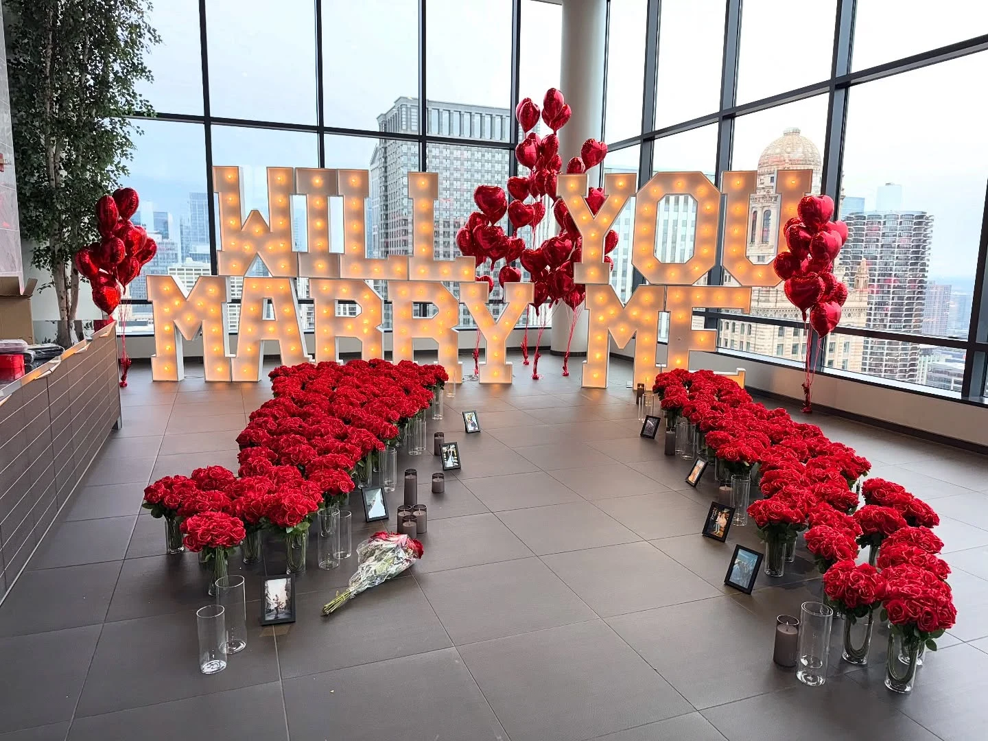 Chicago proposal package with giant Will You Marry Me marquee letters, rows of red rose bouquets, and the downtown skyline through floor-to-ceiling windows