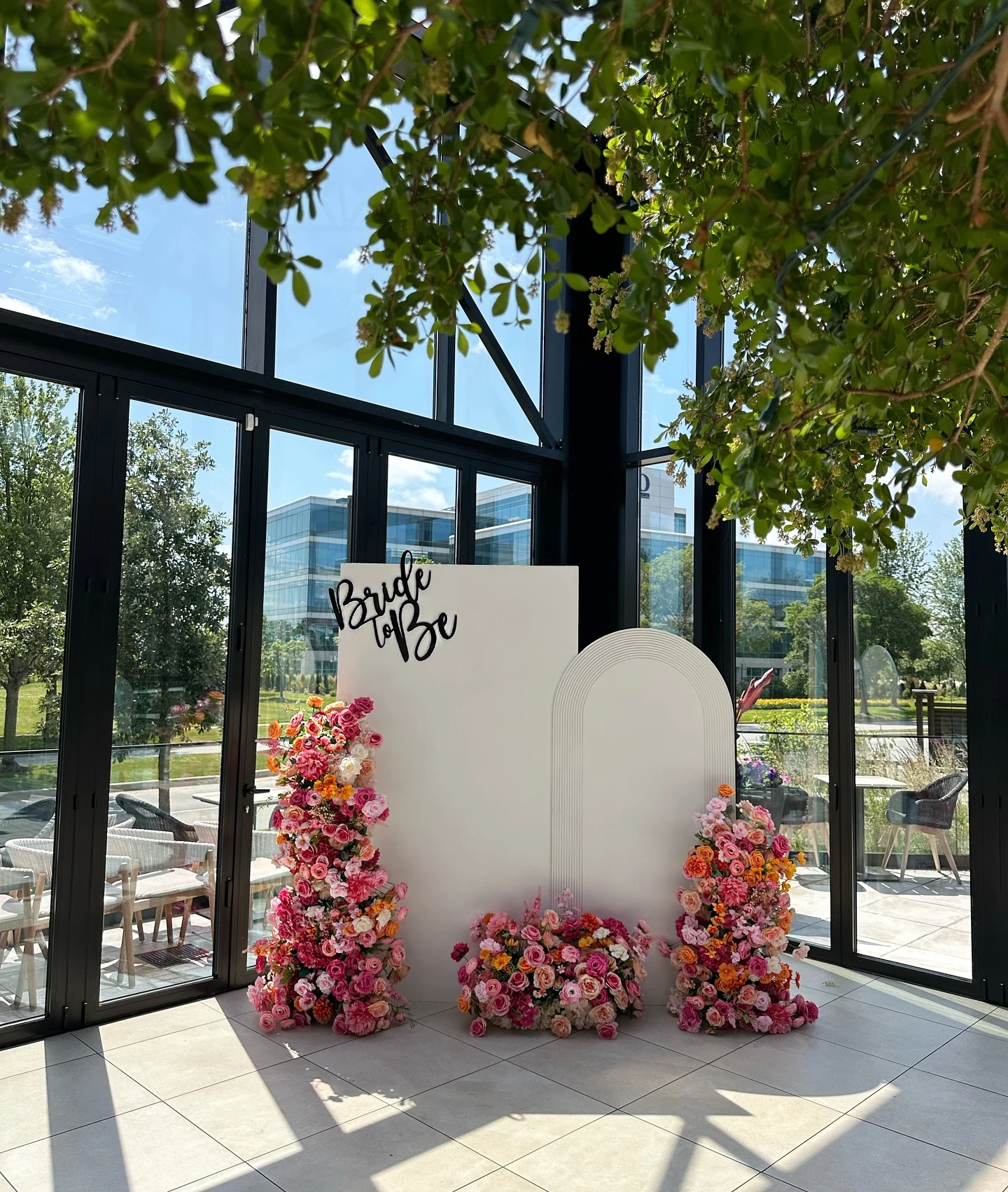 Backdrop rental in Chicago — white Bride to Be bridal shower backdrop with pink, coral and orange floral arrangements in a bright glass-walled room