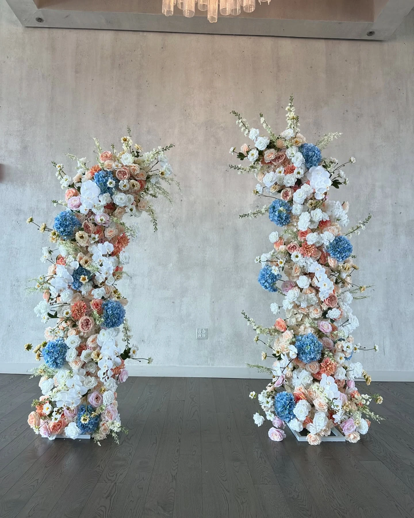Floral pillar rental in Chicago — a new pair of freestanding floral pillars in white roses, blue hydrangeas and peach dahlias standing on a dark wood floor