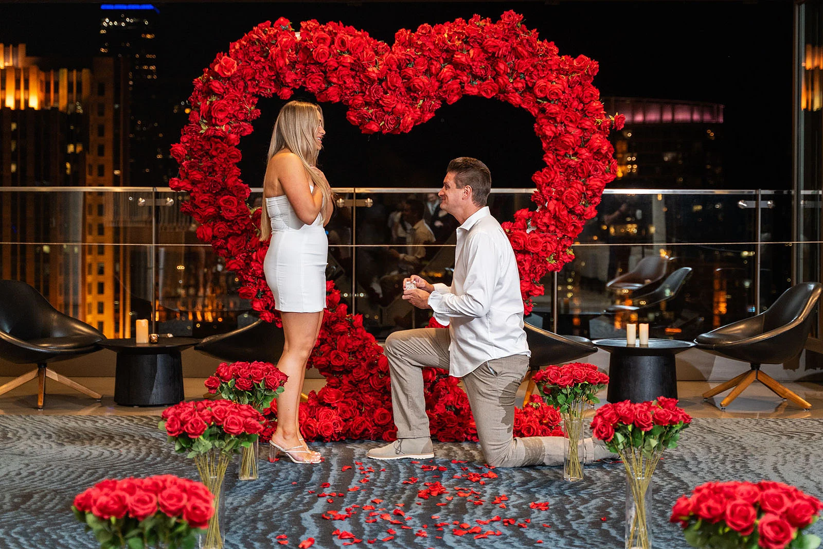 Chicago proposal package at The Lighthouse at 303 — man proposing on one knee inside a red rose heart arch at night, rose bouquets and petals at its base, city lights through the glass